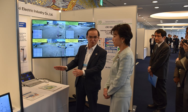 ASTAP-31のOKI展示ブースを見学される佐藤総務副大臣(中央)、左は、説明をする経営基盤本部政策調査部 千村理事