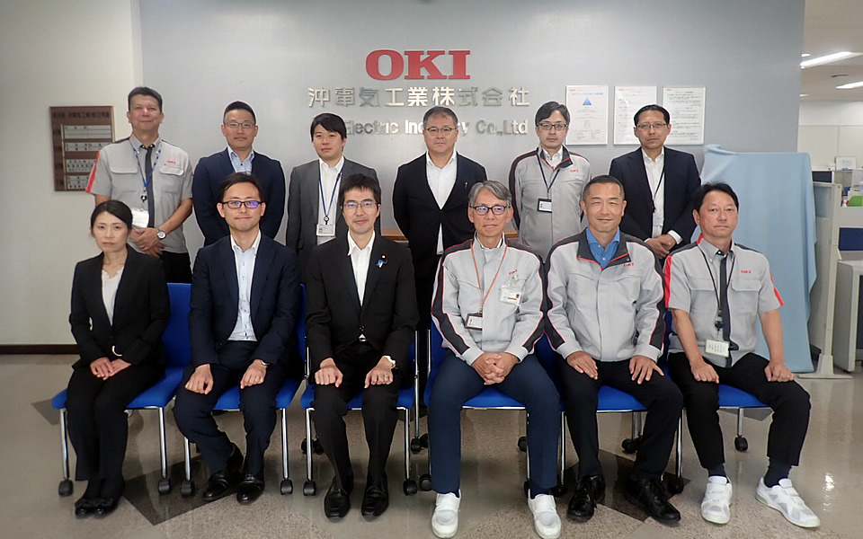 OKI沼津工場にて記念撮影。高見防衛大臣補佐官(前列左3人目)、三橋秘書官(前列左2人目)、櫛田さん(前列左1人目)、安岡さん(後列左2人目)、小原さん(後列左3人目)、田之上さん(後列左4人目)OKI:加藤上席執行役員(前列左4人目)、本杉執行役員(前列左5人目)、海蓋工場長(前列左6人目)